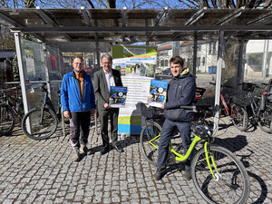 Drei Personen stehen vor den Fahrradständern am Landratsamt Fürstenfeldbruck und halten Plakate in der Hand.