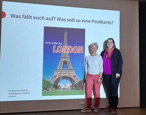 Zwei Frauen stehen rechts nebeneiner Leinwand, auf dem ein Bild des Eifelturms zu sehen ist.